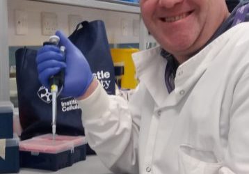 Male researcher, sat next to a lab bench. He is wearing a white lab coat and blue latex gloves and holding a pipette.