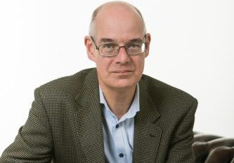 Male sat on a brown leather sofa, draping his hands over the side. He is wearing a pale blue shirt and brown suit style jacket.