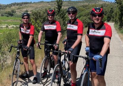 four men wearing the same red and black cycling kit, standing next to their bikes, with blue skies behind them.