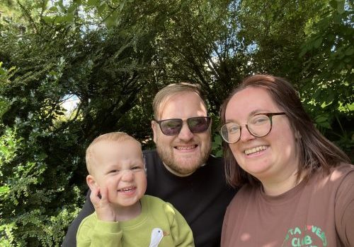 Family selfie of dad, mum and small boym with hedge and trees in the background.