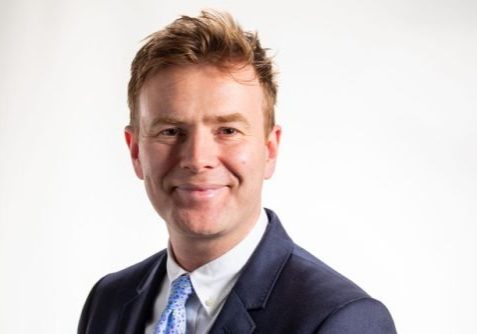 Head and shoulders picture of a man wearing a suit, white shirt and blue tie, smiling for the camera.