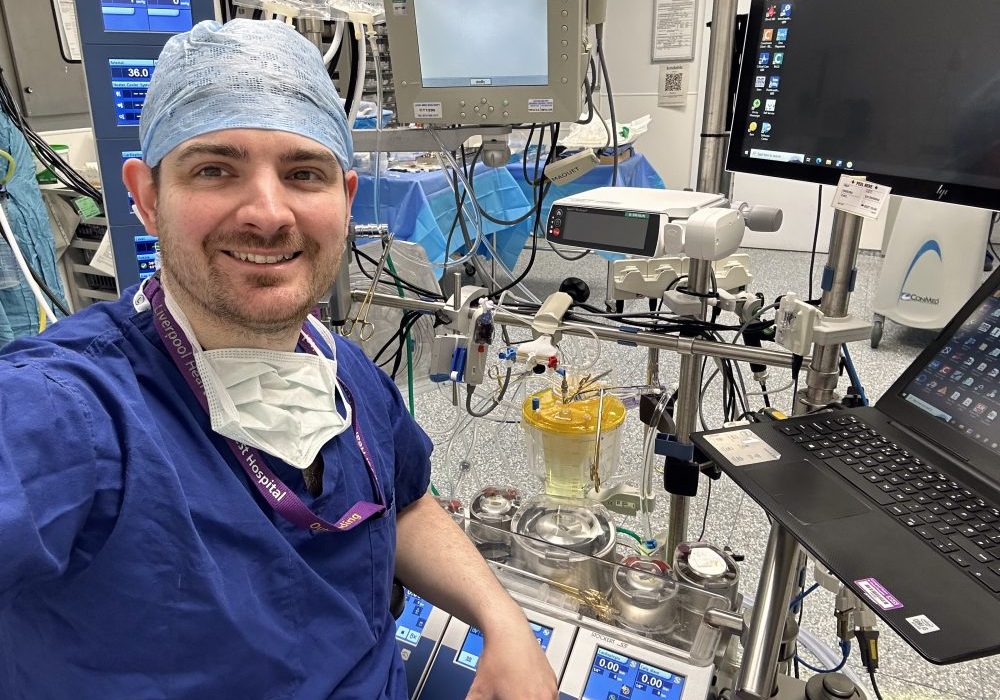 A male, dressed in blue hospital scrubs, in a operating room surrounded by equipment and a computer next to him,