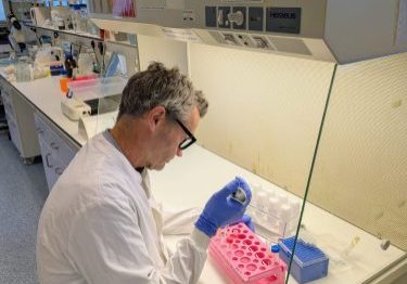 Male researcher working at a bench, wearing blue gloves and white coat, holding a pipette