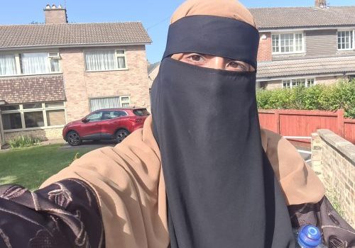 Ummae Kulsoom (1) Close up of an Asian female, wearing a tan colour and black burka. Houses in the background