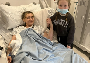 Ellie-Mae Steers Female in hospital bed with young girl standing next to the bed holding her hand.